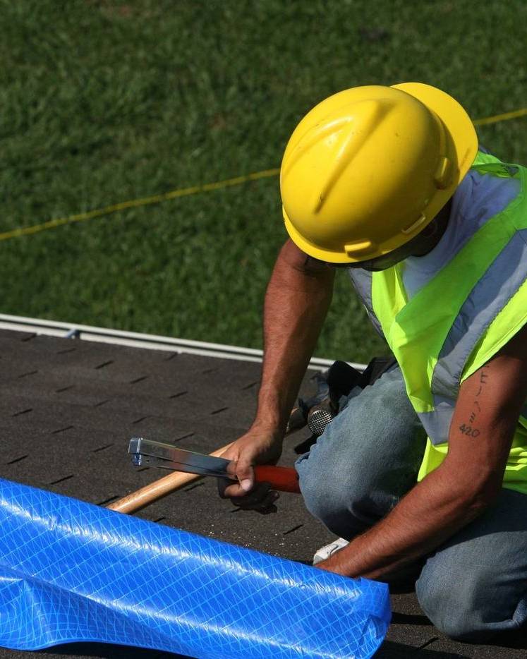 A worker fastens a blue tarp over a leaking roof